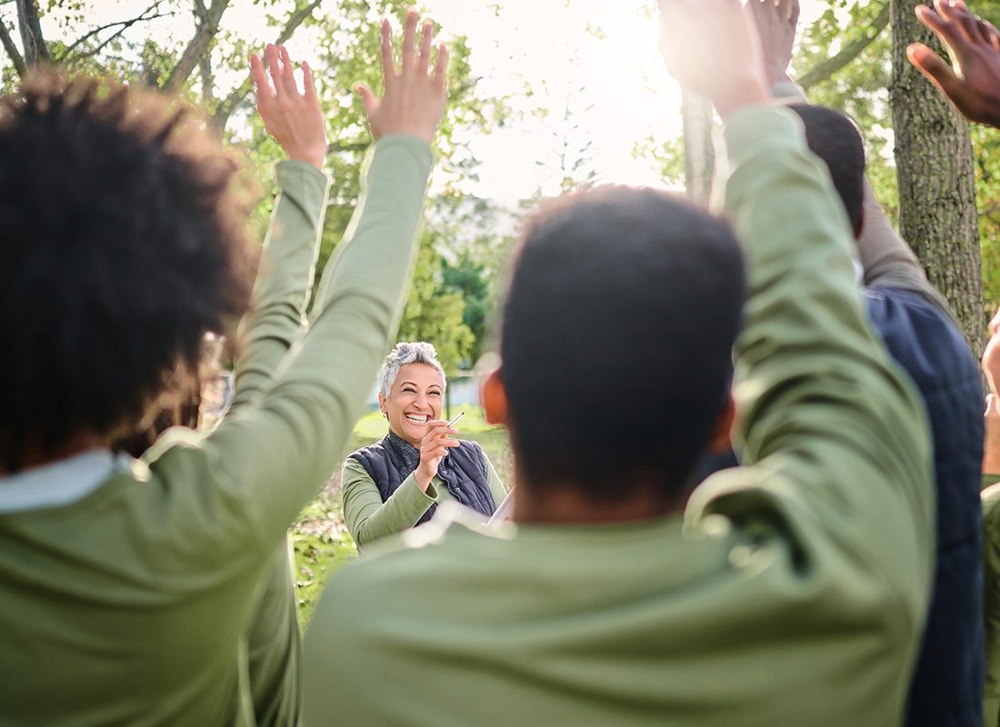 About Our Agency - Cheerful Elderly Woman Holding Up a Pen While Standing in Front of a Group of People Waving Their Hands in the Air Outside in During a Volunteer Event in a Park