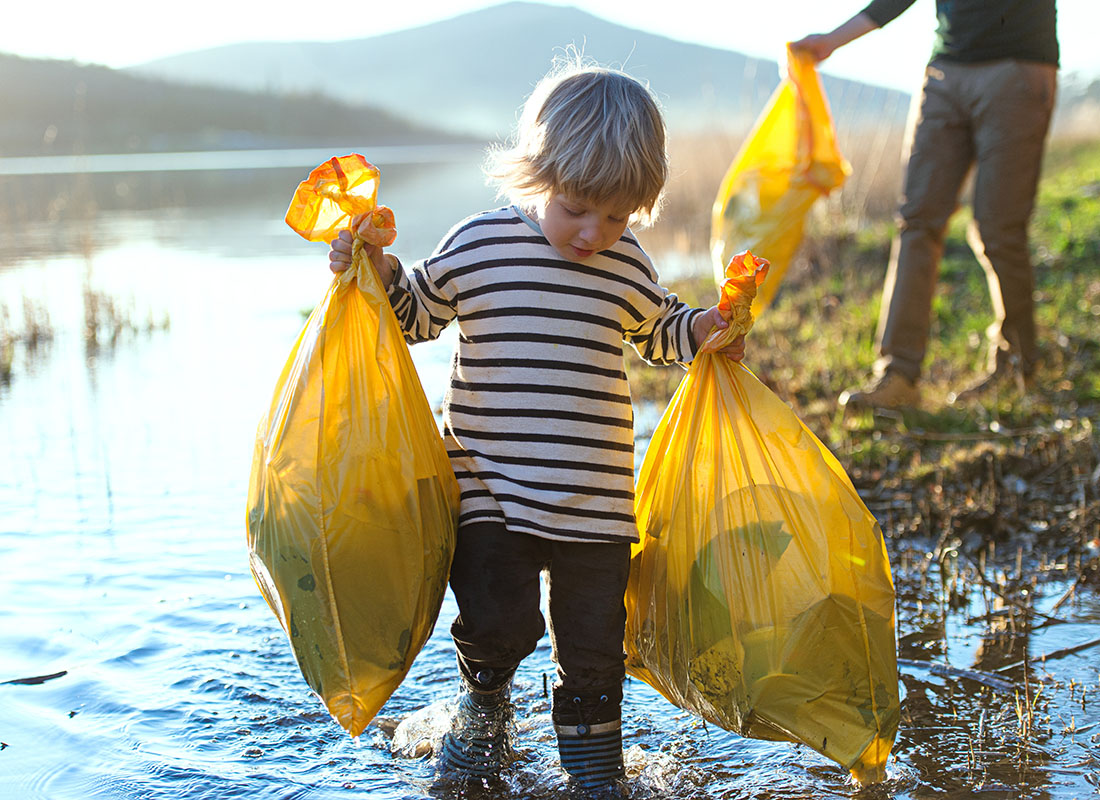 Contact - Closeup View of a Young Boy Holding Up Two Yellow Trash Bags While Walking Along the Edge of a Lake Doing Volunteer Work with his Father